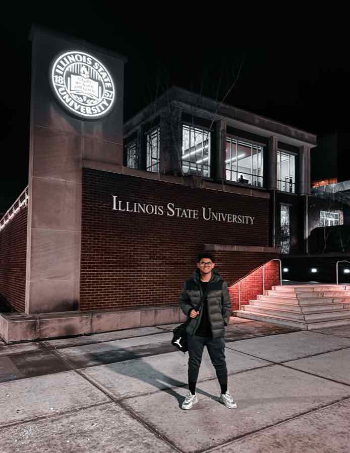 Salman smiling at night in front of a brick building with the lit Illinois State University seal and lettering.