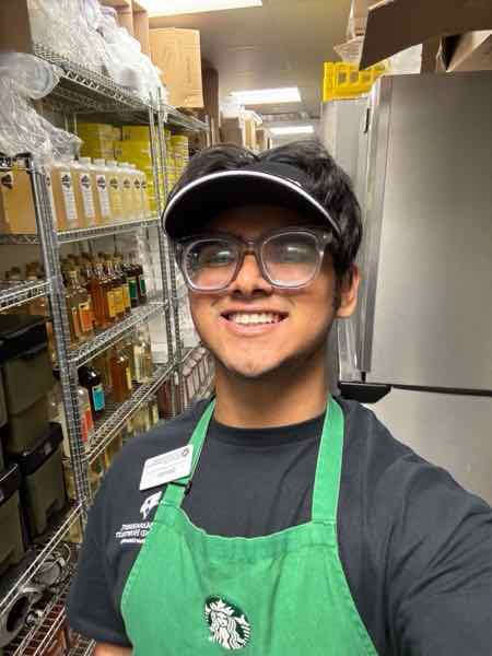Salman smiling in a green Starbucks apron and visor in the stockroom.